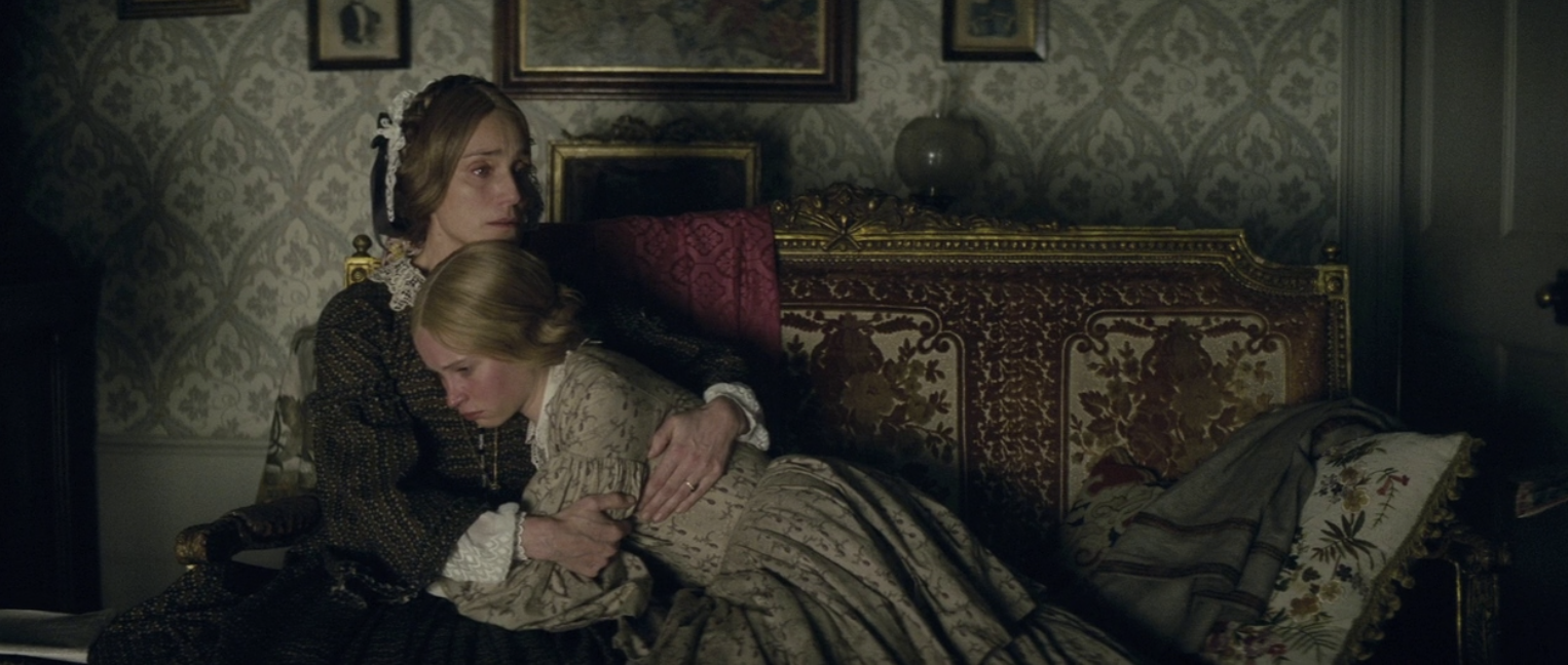 Two women in period dress embracing on an ornate couch